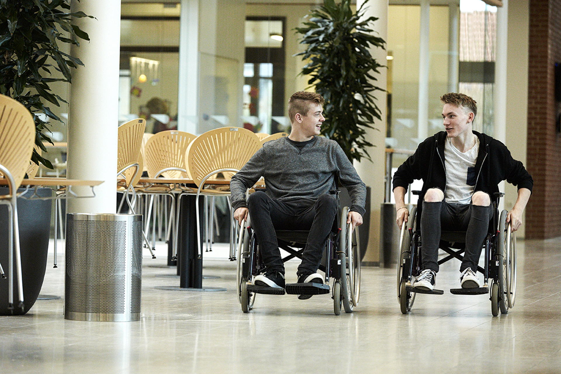 To unge fyre sidder i hver sin kørestol i skolens fællesrum. De er iført mørkt tøj og kigger på hinanden, mens deres hænder er på hjulene af kørestolene. 