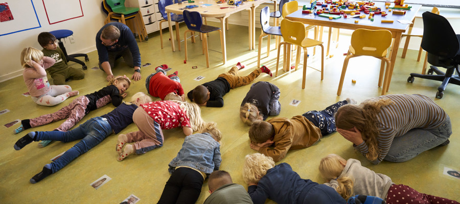 En gruppe børnehavebørn ligger på maven på gulvet på stuen, hvor de leger en leg sammen med to voksne. Gulvet er grønt, og alle børnene er iført farvestrålende tøj.